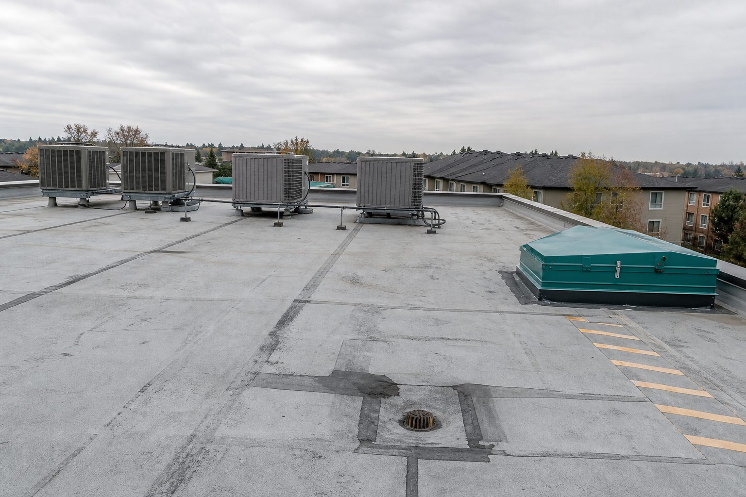 Low slope apartment roof with patched membrane seams, a roof drain, and rooftop HVAC units under grey sky